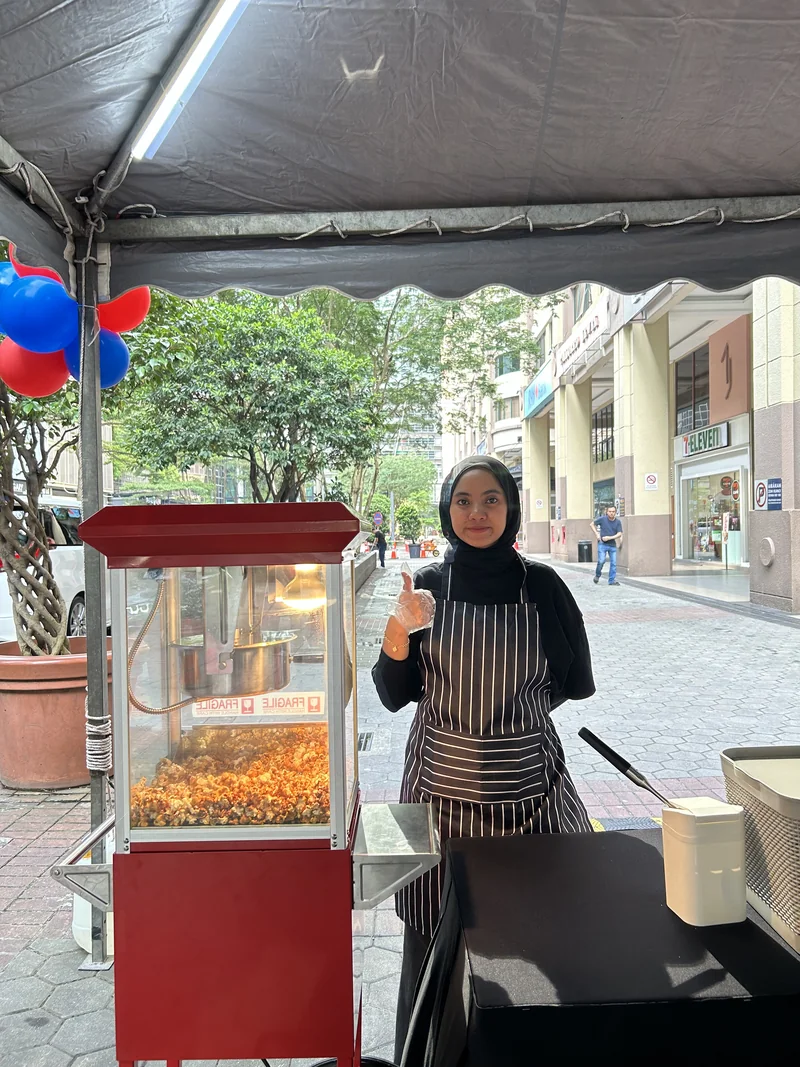 An operator serving popcorn from a red popcorn machine under an event tent