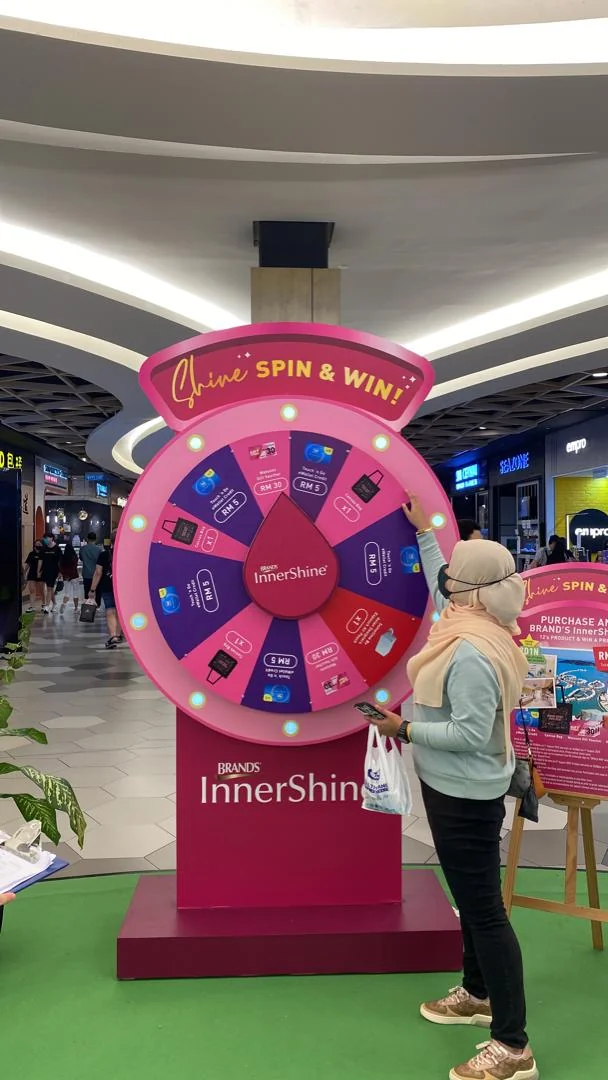 Guest beside a pink BRAND'S InnerShine prize wheel at a shopping-centre event